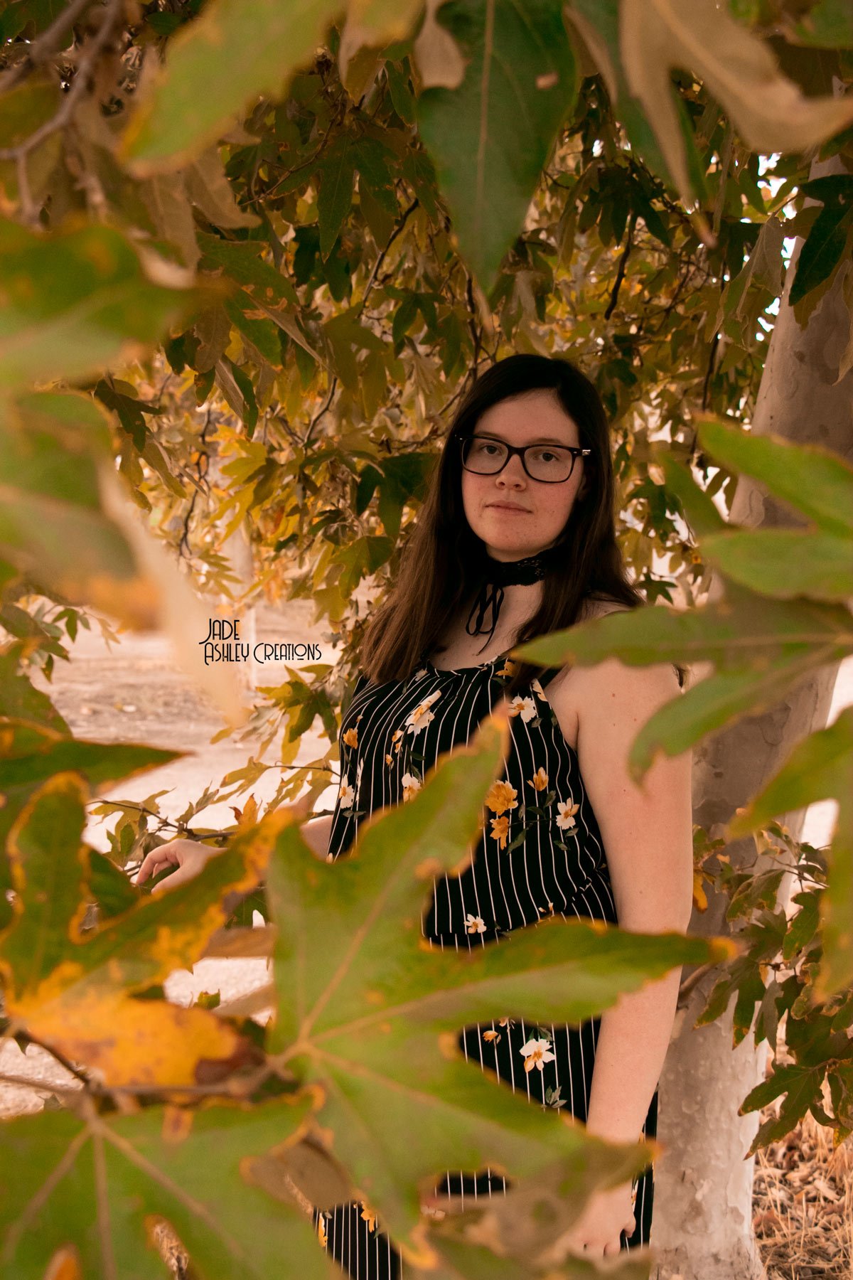 A young woman with dark hair and glasses standing amidst autumn leaves on a tree, wearing a black dress with white and yellow floral patterns and a black choker with bow.