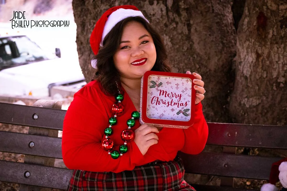 Woman dressed in red Christmas attire, wearing a Santa hat and holiday necklace, holding a box with 'Merry Christmas' written on it, sitting on a park bench outdoors.