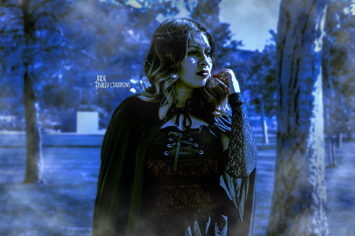 A woman with dark hair and fairy lights in her hair, wearing dark makeup and a gothic outfit with lace sleeves, standing outdoors near trees with a blue-tinted filter applied to the photo.