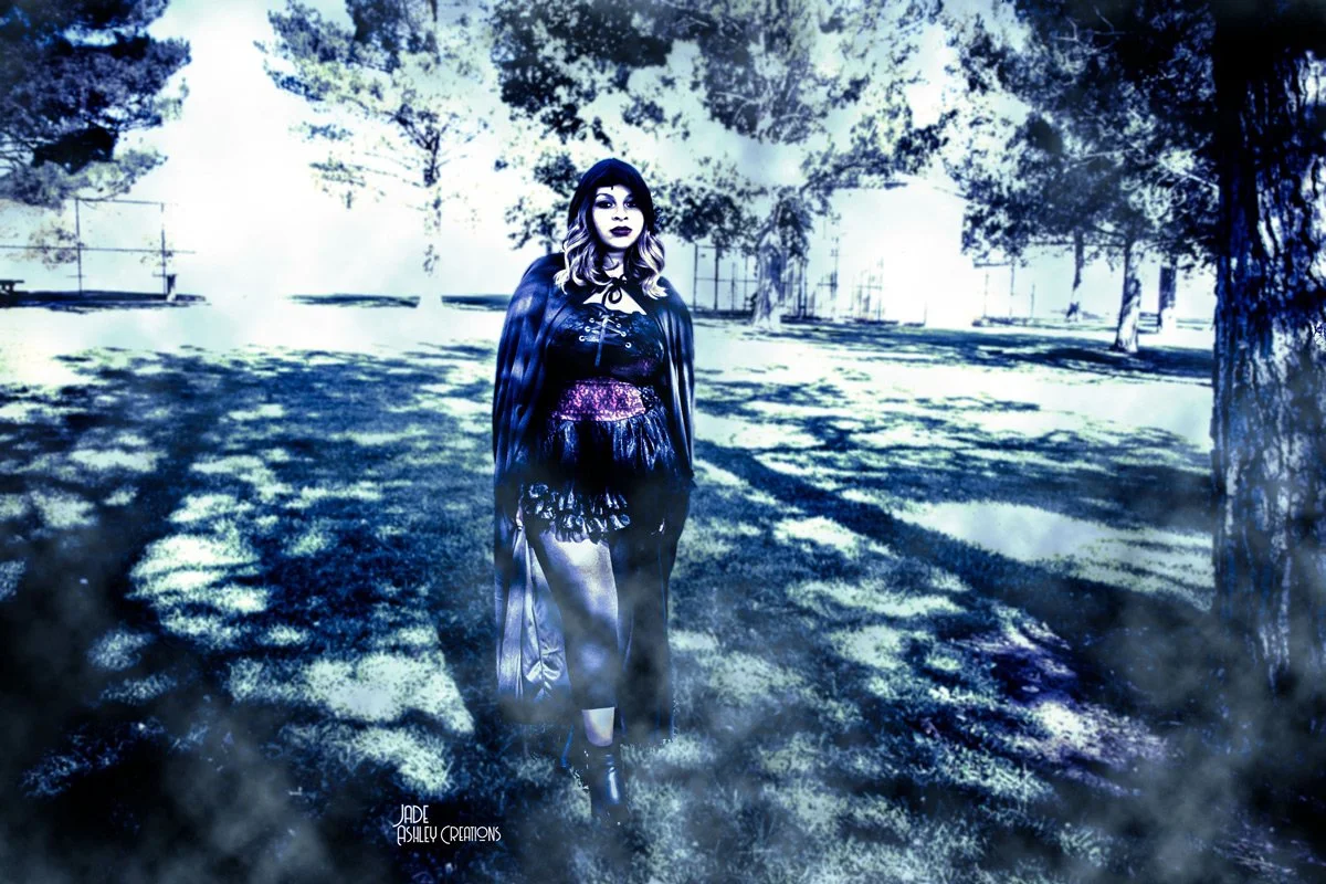 A woman dressed in dark, Gothic-style clothing standing in an outdoor park with trees, benches, and a fence in the background. The image has a surreal, high-contrast, almost monochromatic blue and black tone.