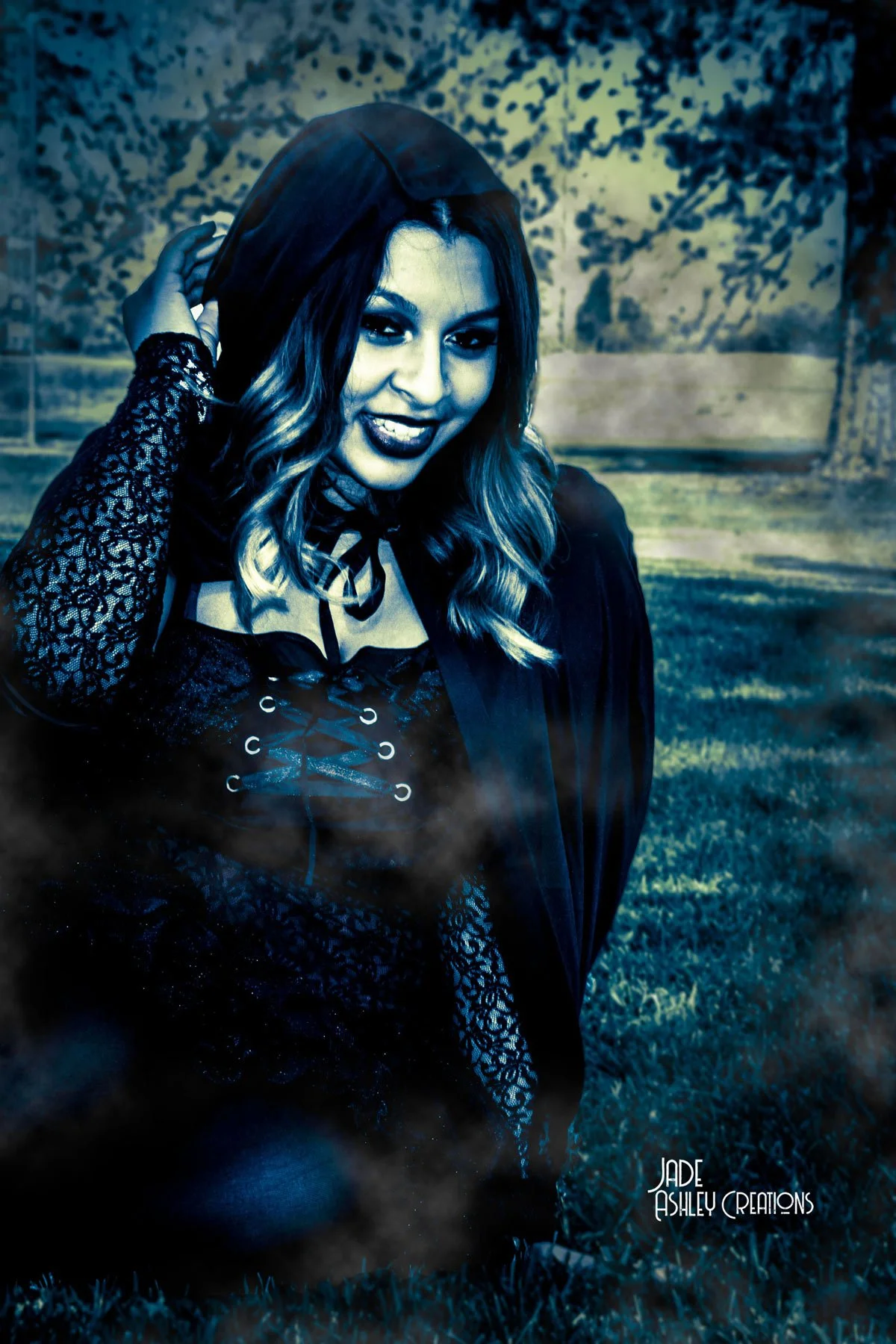 Young woman in Gothic attire outdoors, smiling and holding her hood, with a dark, mysterious atmosphere and fog.