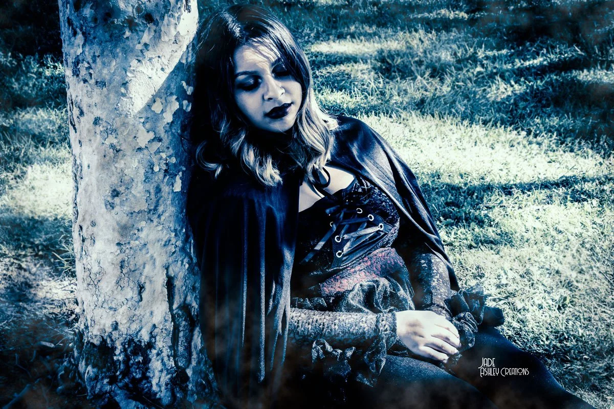 A woman with dark makeup, blonde hair with dark roots, wearing gothic clothing, sitting against a tree in a grassy area, with her eyes closed and a somber expression.