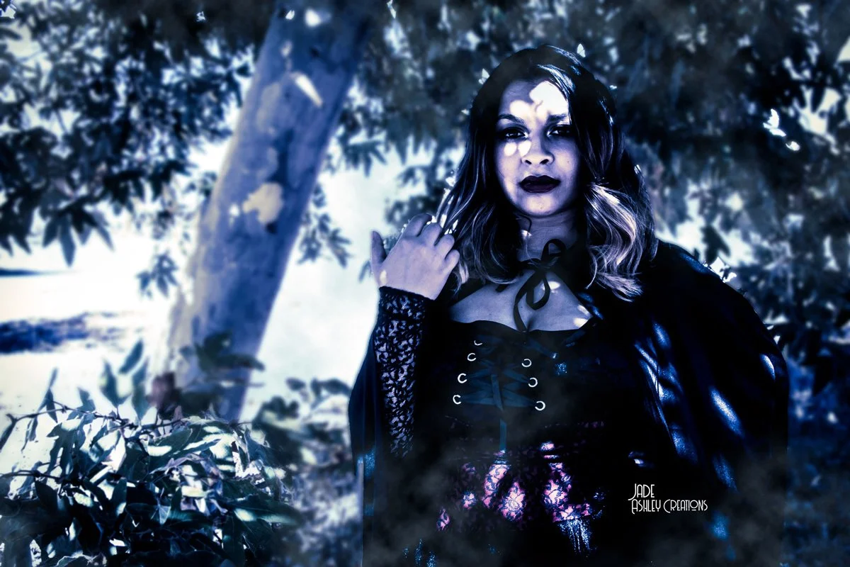 A woman dressed in black gothic clothing with dark makeup poses in front of a tree and leafy background, with dramatic lighting creating shadows on her face.