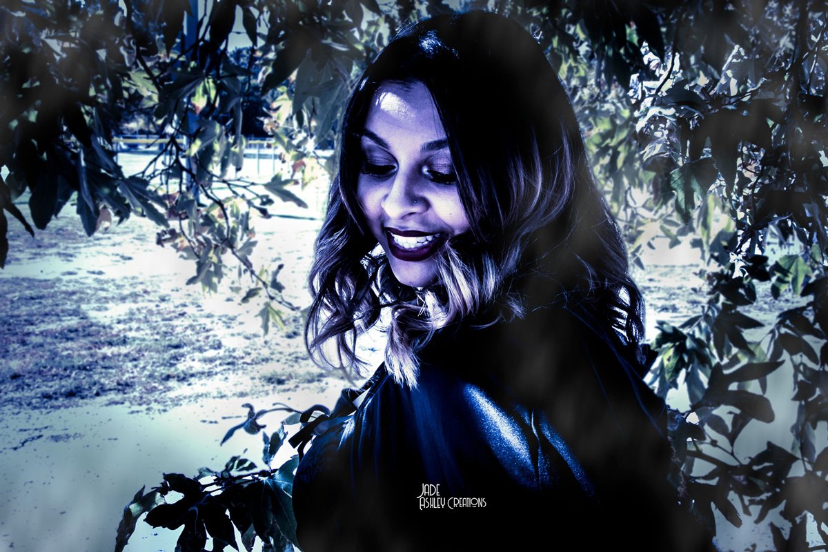 A woman with dark hair and makeup standing outdoors in front of leafy trees, smiling and looking downward