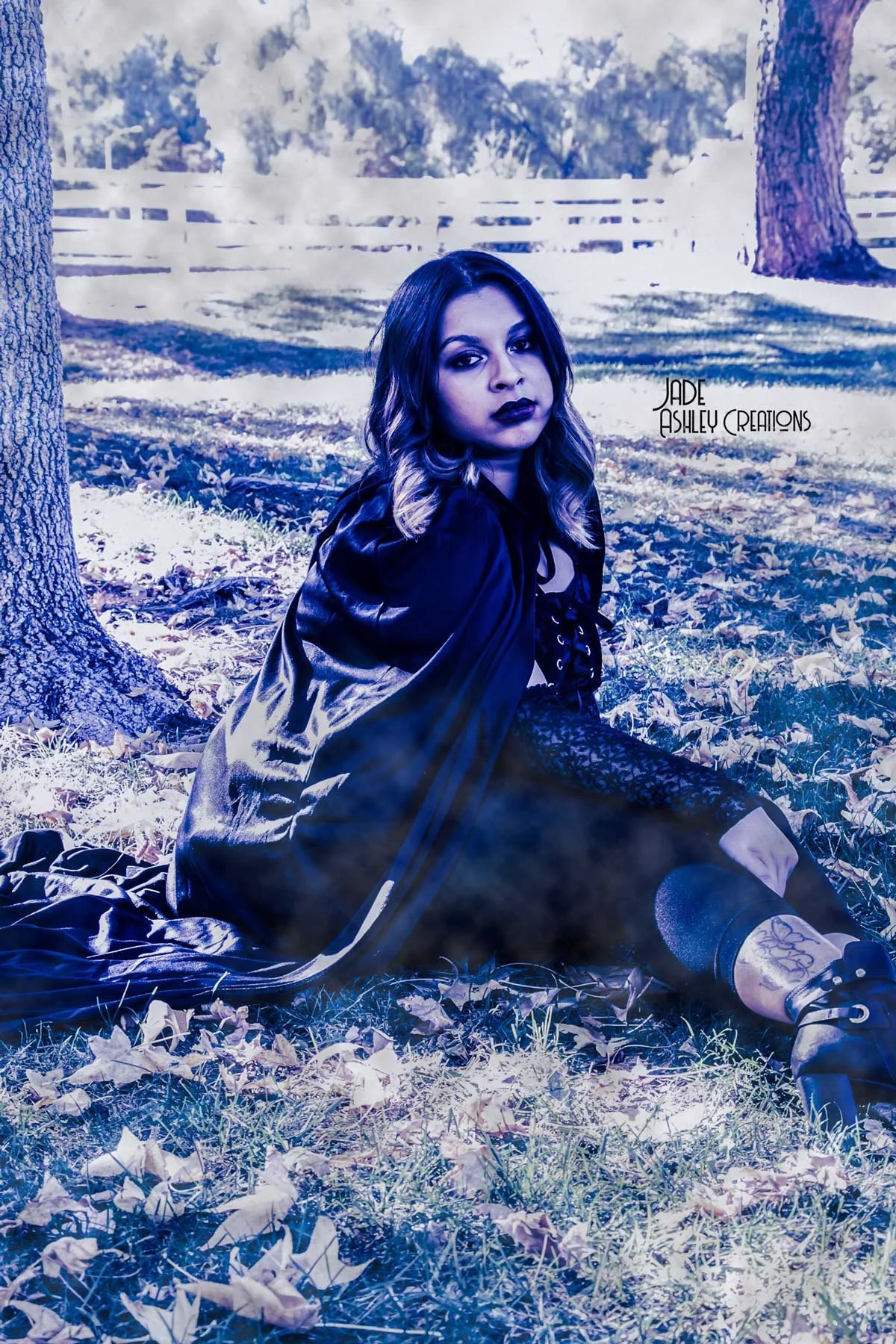 A woman with dark lipstick and wavy hair sitting on the ground beside a tree in a park, with a white fence and trees in the background, and the text 'Jade Ashley Creations' on the right side of the image.