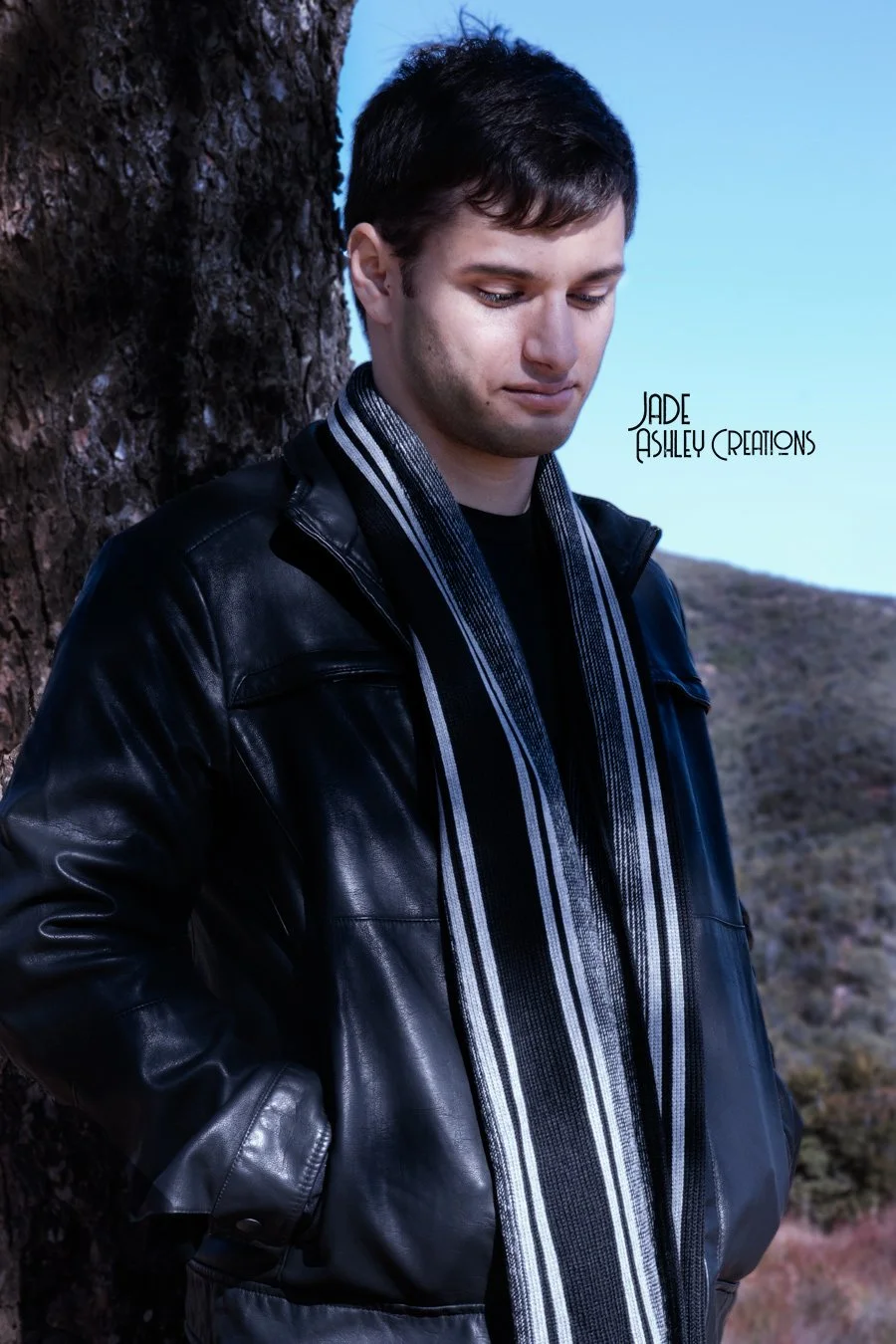 Young man with dark hair, wearing a black leather jacket and a striped scarf, standing outdoors next to a tree with a hill in the background.