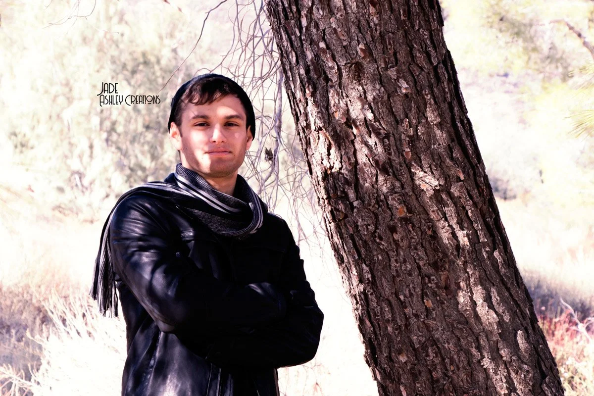 A young man with dark hair, wearing a black leather jacket and a black scarf, standing outdoors next to a large tree with a rough textured bark. He has his arms crossed and is looking at the camera.
