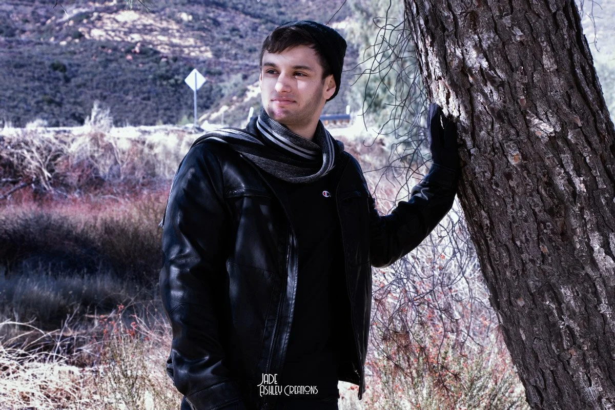 A young man standing outdoors next to a tree, wearing a black leather jacket, a black and gray scarf, and a black beanie, with a scenic hilly landscape in the background.