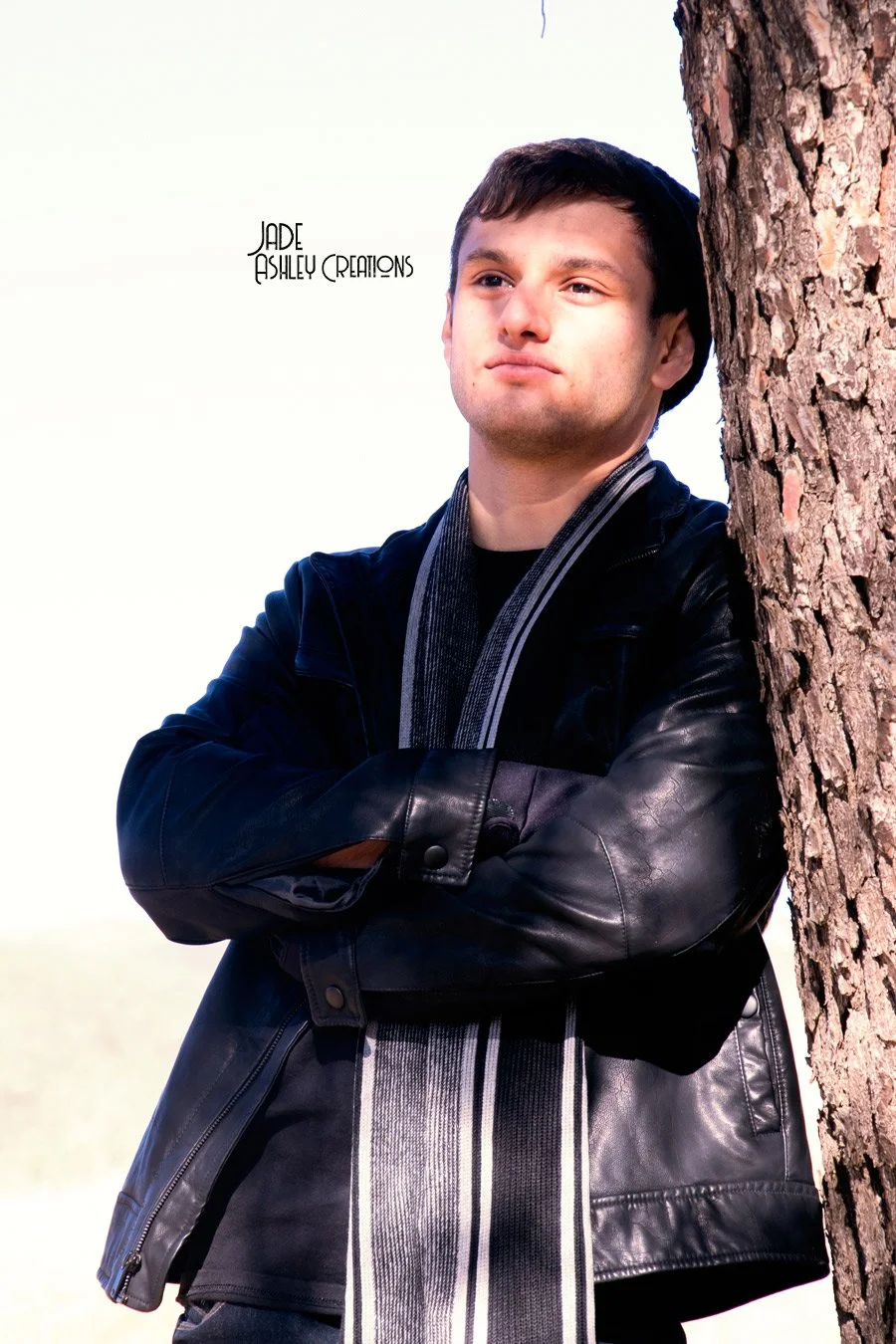 Young man in a black leather jacket and striped scarf leaning against a tree with his arms crossed, outdoors on a bright day.