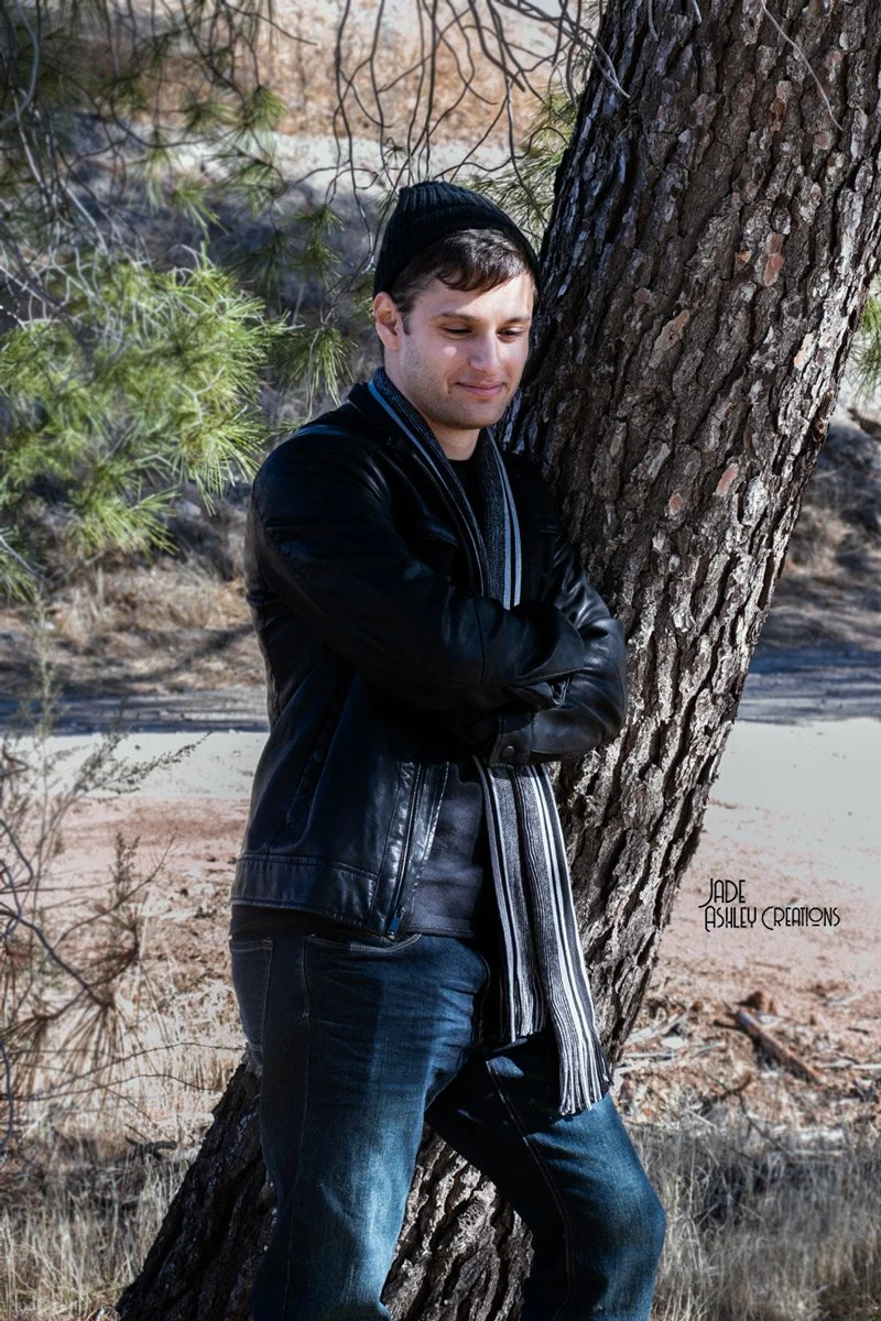 A young man with short brown hair wearing a black beanie, black leather jacket, striped scarf, and jeans, leaning against a large tree outdoors in a wooded area.