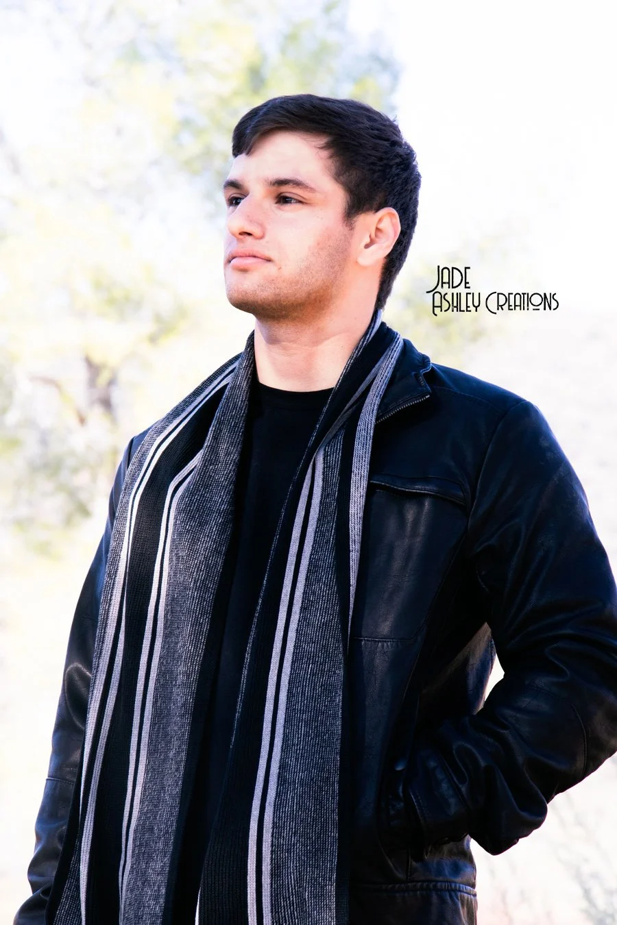 Portrait of a young man with dark hair, wearing a black leather jacket and striped scarf, outdoors with blurred trees in the background.