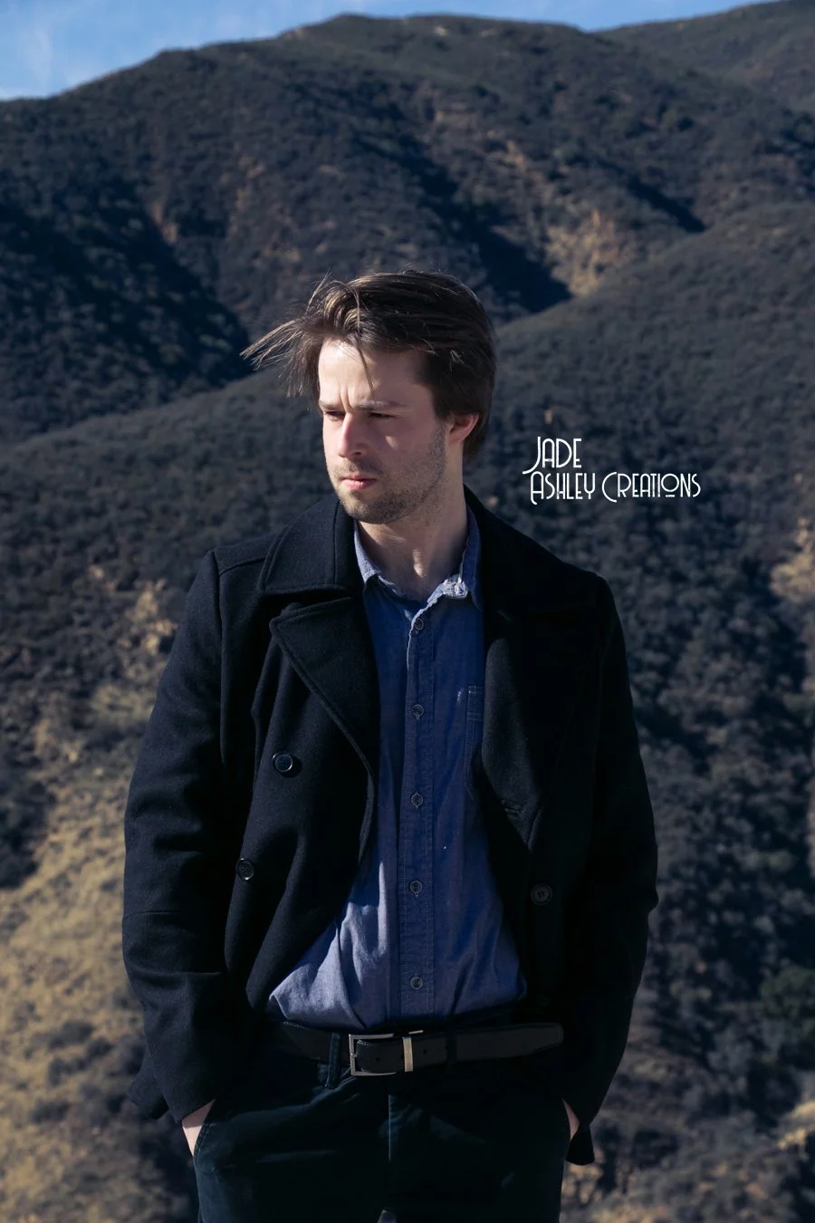 A man standing outdoors in front of mountainous terrain with dry bushes, wearing a dark coat over a blue shirt, with hands in pockets and looking to the side.
