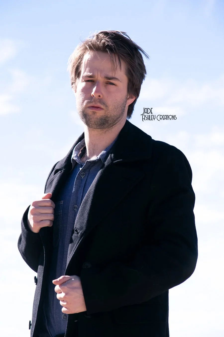 A man standing outdoors under a blue sky with clouds, wearing a dark coat and a collared shirt, looking into the camera with a serious expression.