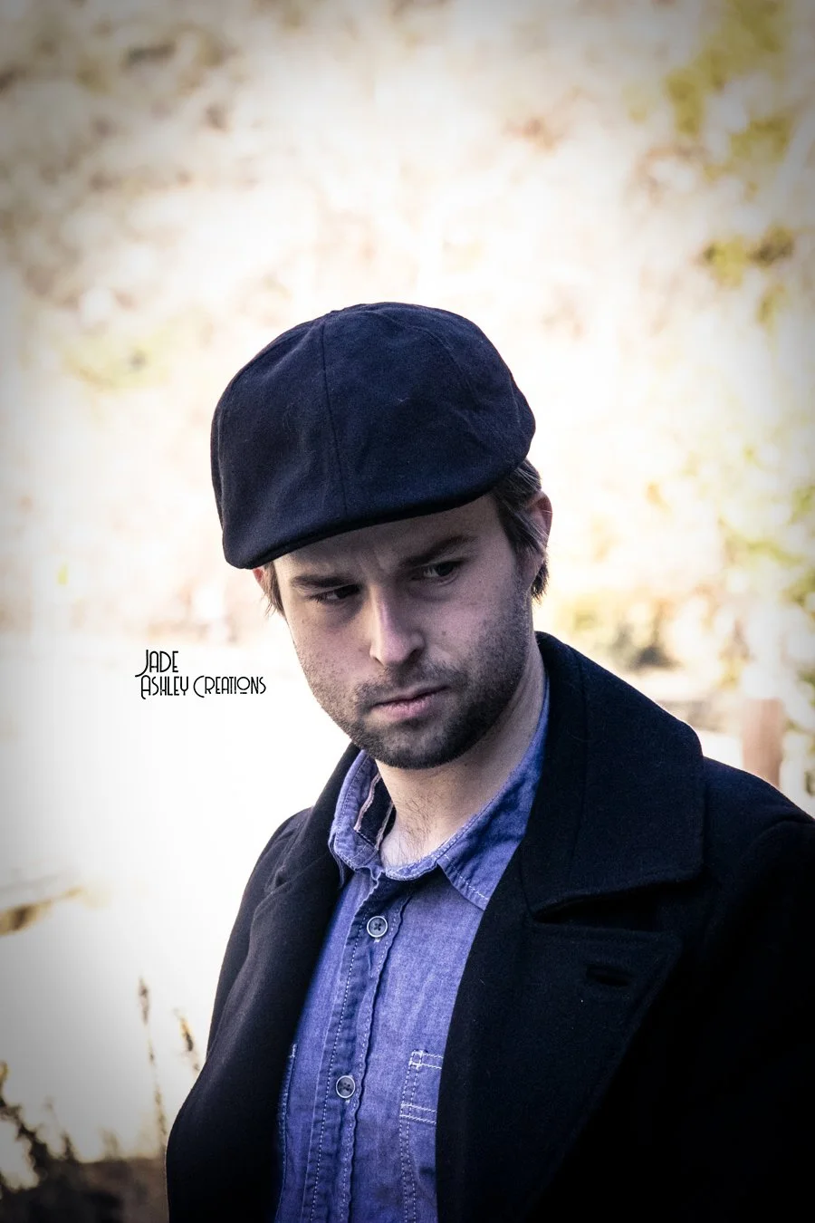A young man with light skin, dark hair, and facial hair wearing a black beret, a dark coat, and a blue button-up shirt outdoors, looking slightly to the side.