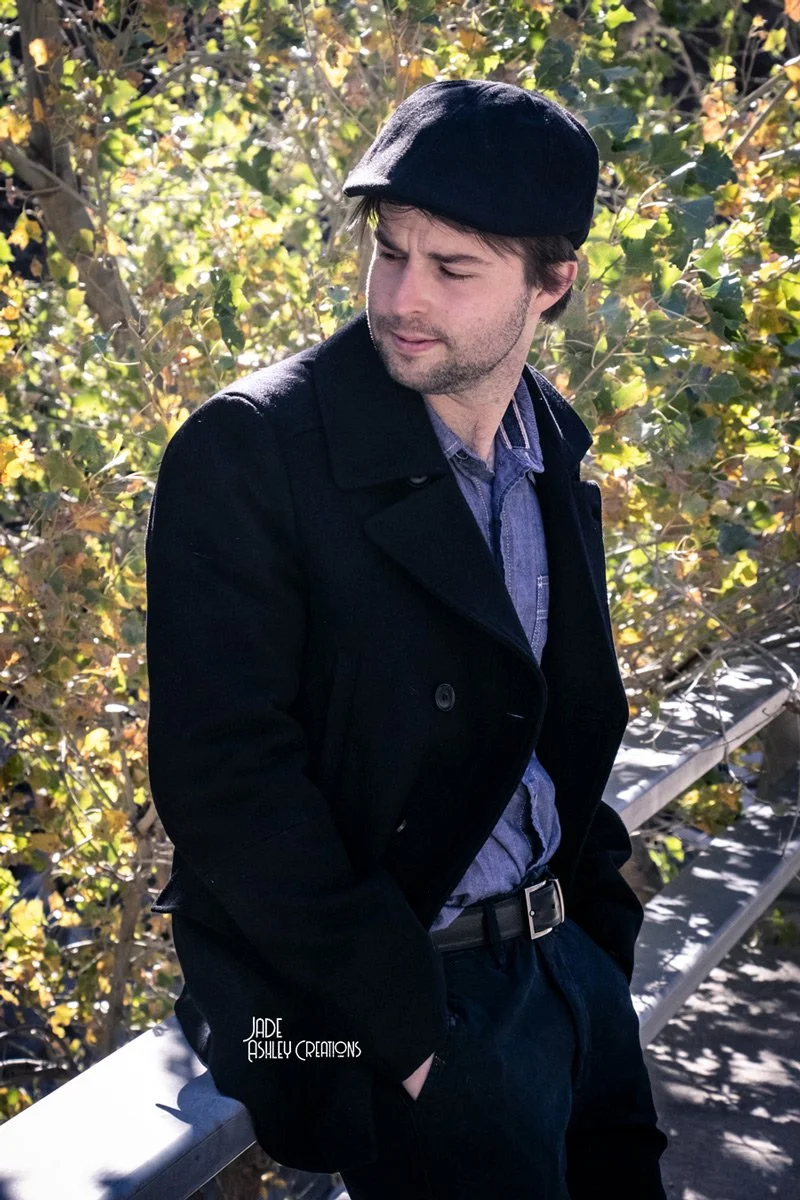 A man with dark hair and a beard wearing a black coat, a blue shirt, and a black hat, standing outdoors on a bridge or railing with foliage and trees in the background.