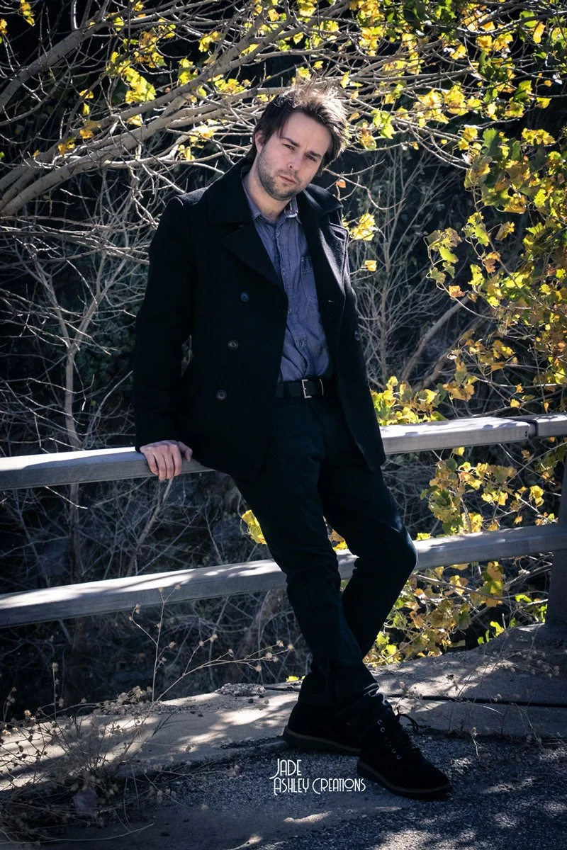 A young man with dark hair and a beard leaning against a metal railing outdoors, surrounded by leafless trees and some green leaves, wearing a dark coat and black pants.