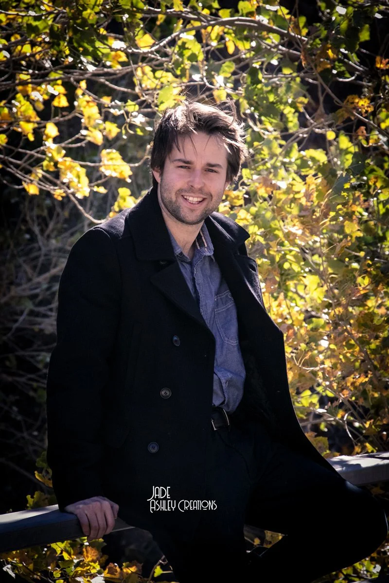A man with brown hair and a beard smiling outdoors, sitting on a railing with autumn foliage in the background.