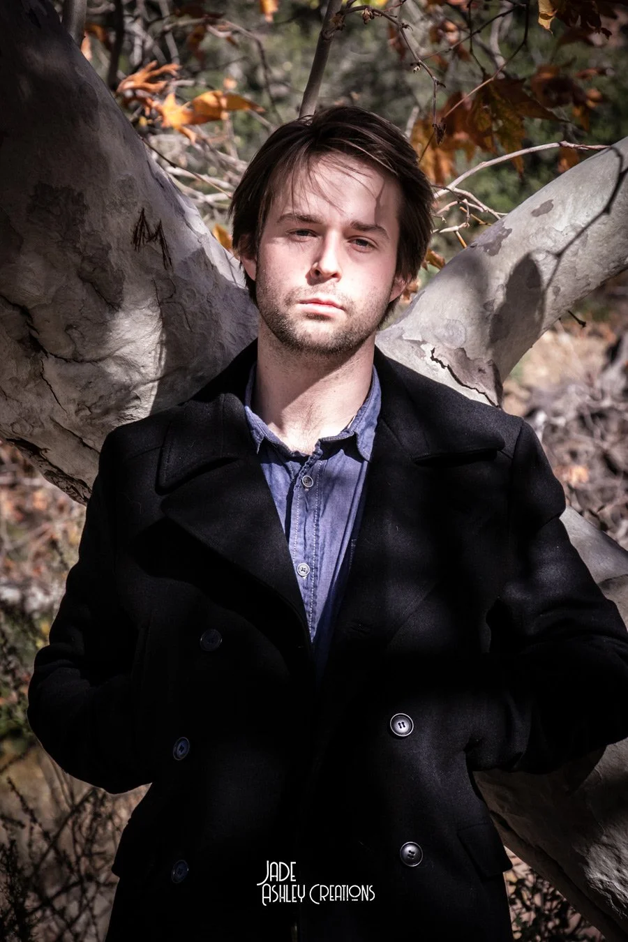 A young man with dark hair and light skin leaning against a tree outdoors, wearing a dark coat over a blue shirt, with a neutral expression, surrounded by autumn leaves.