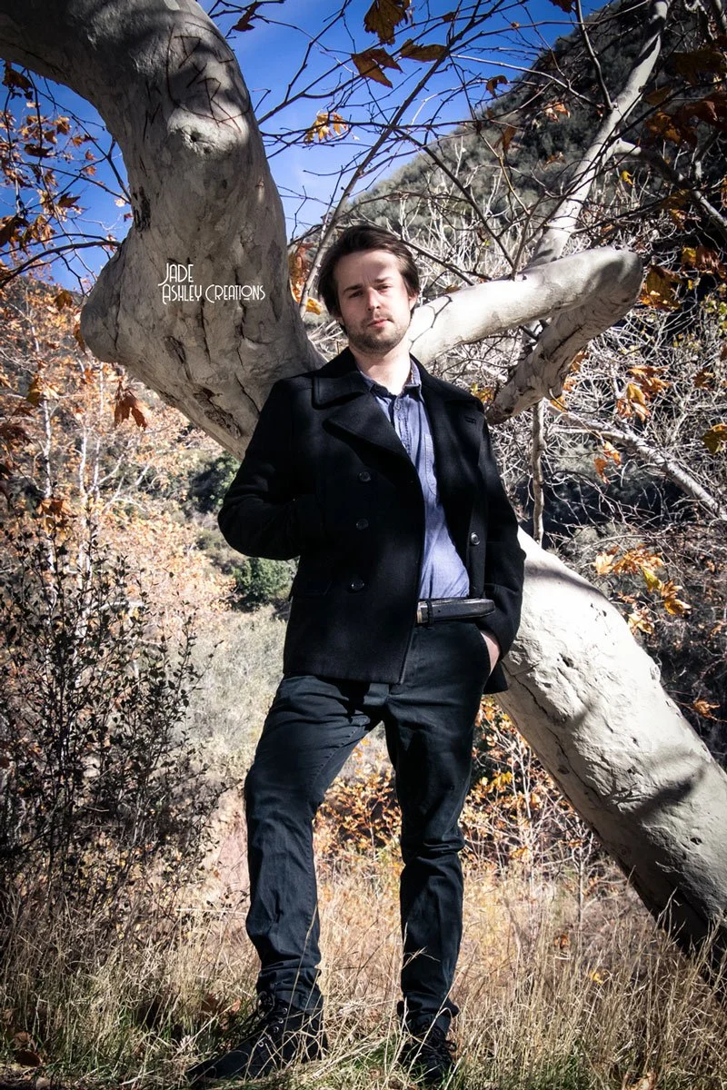 A young man with dark hair and a beard standing in front of a large, curved tree with a rugged, gray bark and brown autumn leaves. The background features a mountain slope and a clear blue sky.
