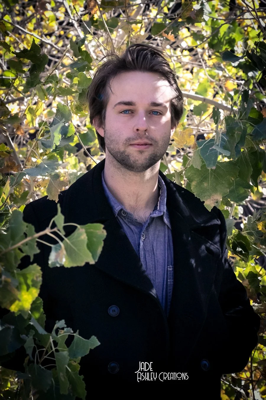 A young man with dark hair and blue eyes standing among green leafy branches with sunlight filtering through.