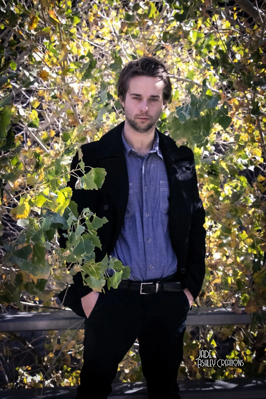A young man with brown hair and a beard standing outdoors in front of leafy green and yellow foliage. He is wearing a blue button-up shirt, black pants, and a black jacket, with hands in his pockets.