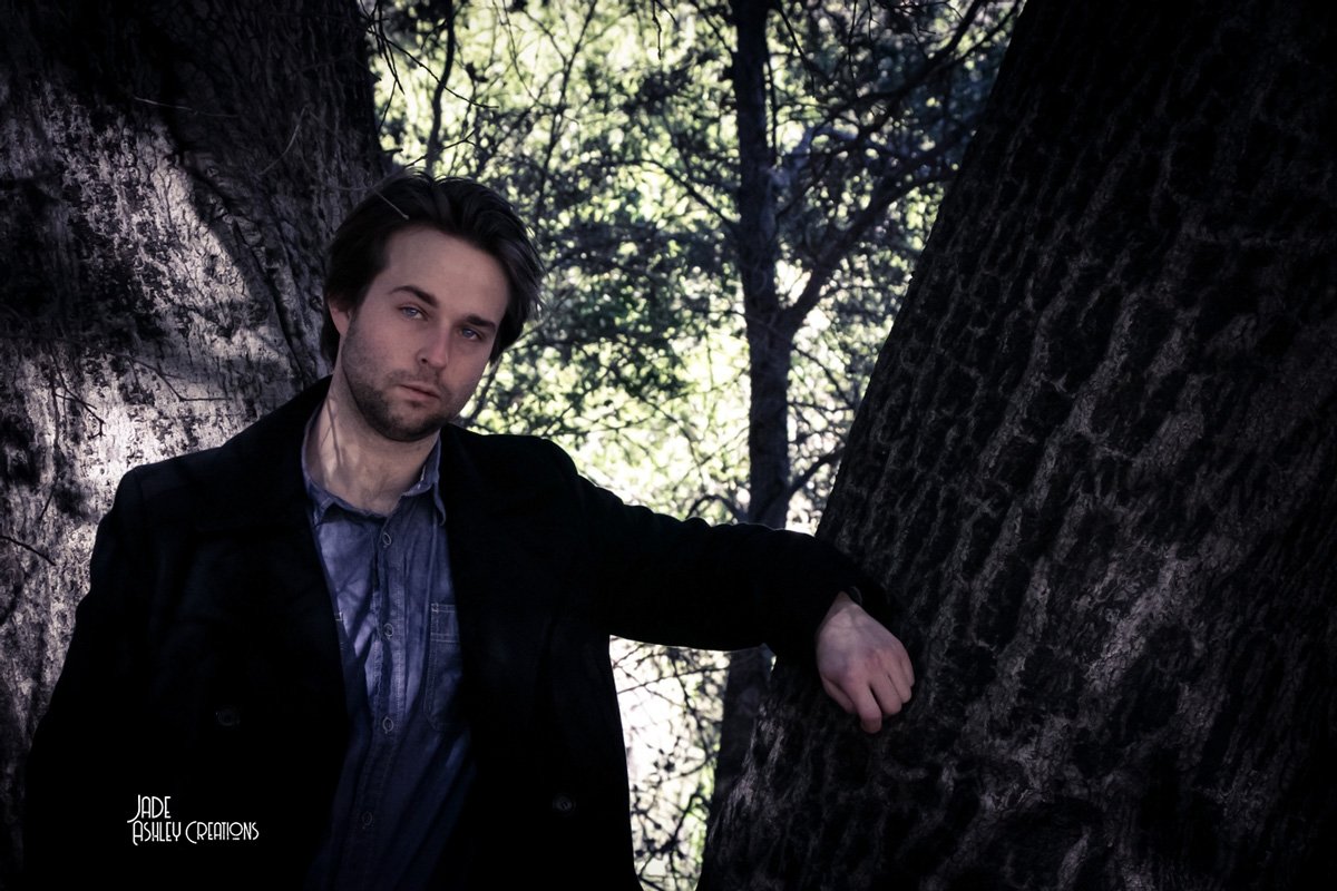 A man with dark hair and a beard leaning against a tree in a wooded area, wearing a dark jacket and blue shirt, with sunlight filtering through the trees in the background.