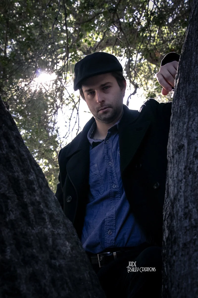 A man with dark hair and facial hair, wearing a black cap and black jacket, leaning against a tree in a wooded area with sunlight filtering through the leaves.