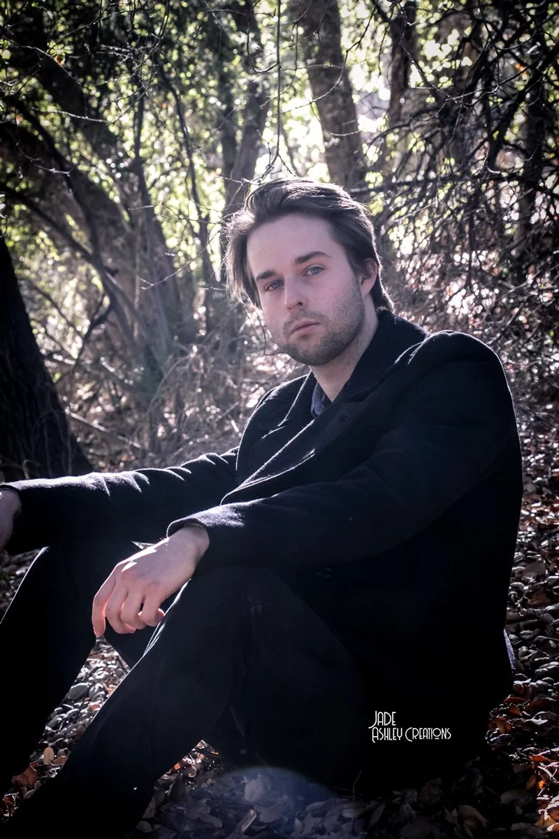 A young man with dark hair and light skin sitting outdoors among trees and fallen leaves, wearing a black jacket and black pants, looking seriously at the camera near sunlight filtering through the branches.