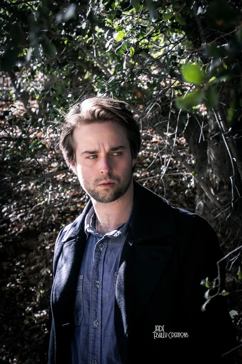 A young man with brown hair and a beard standing outdoors among dense greenery, wearing a dark jacket over a blue button-up shirt, looking to his left with a serious expression.