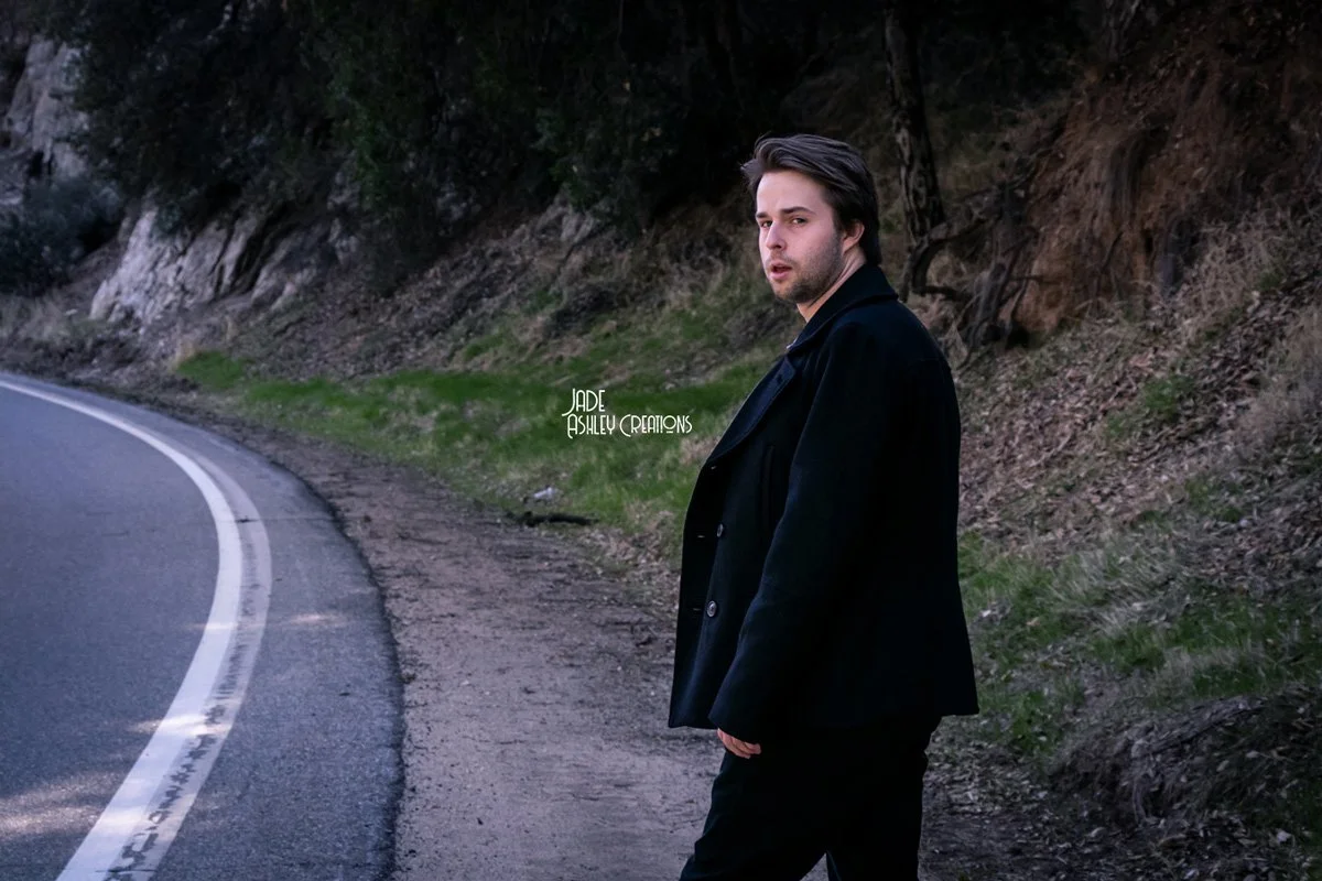 A young man with dark hair and a beard walking on the side of a curved road surrounded by trees and grass during daytime.