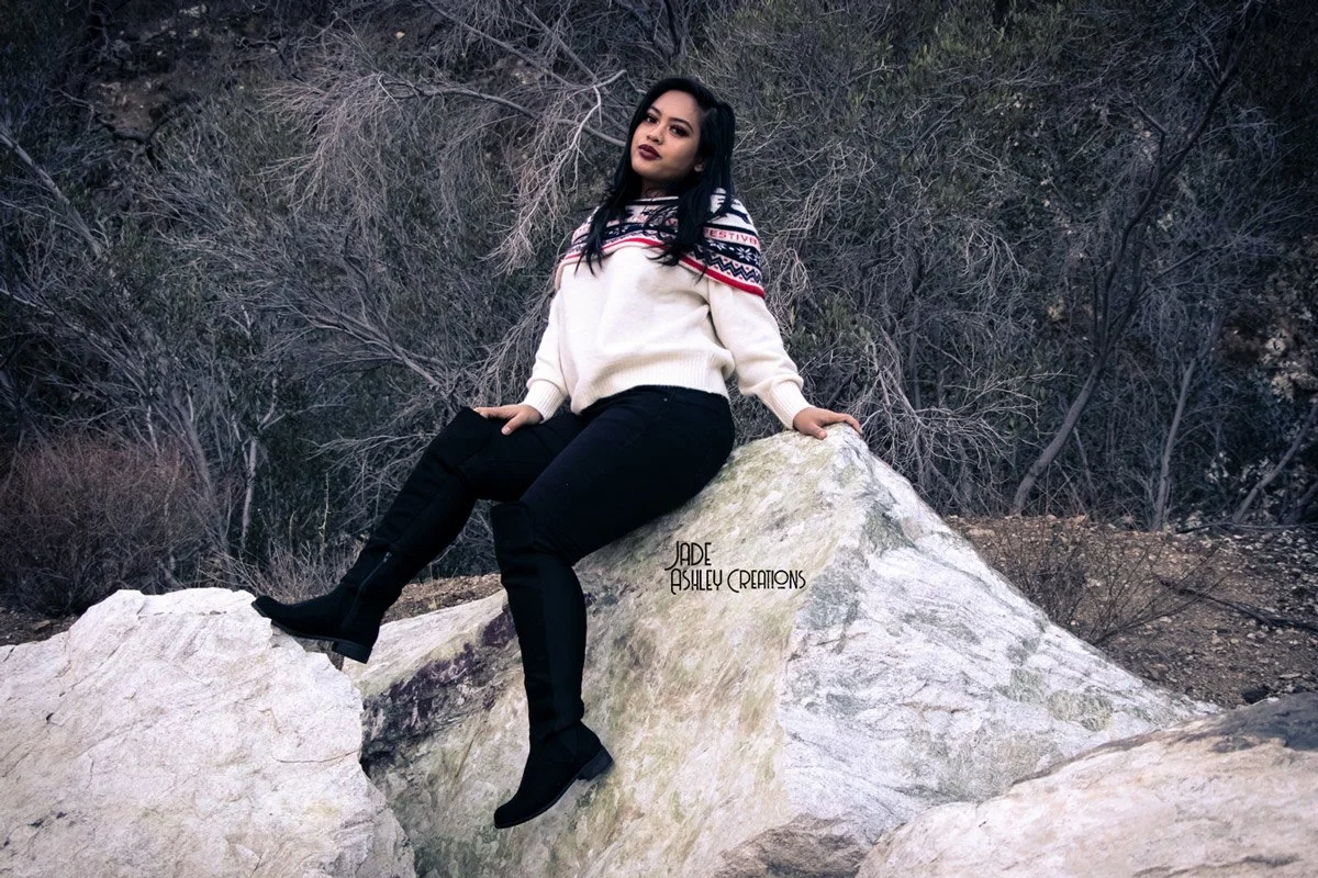 A woman sitting on a large white rock outdoors, surrounded by desert vegetation, wearing a cream sweater with colorful patterns, black pants, and black boots.