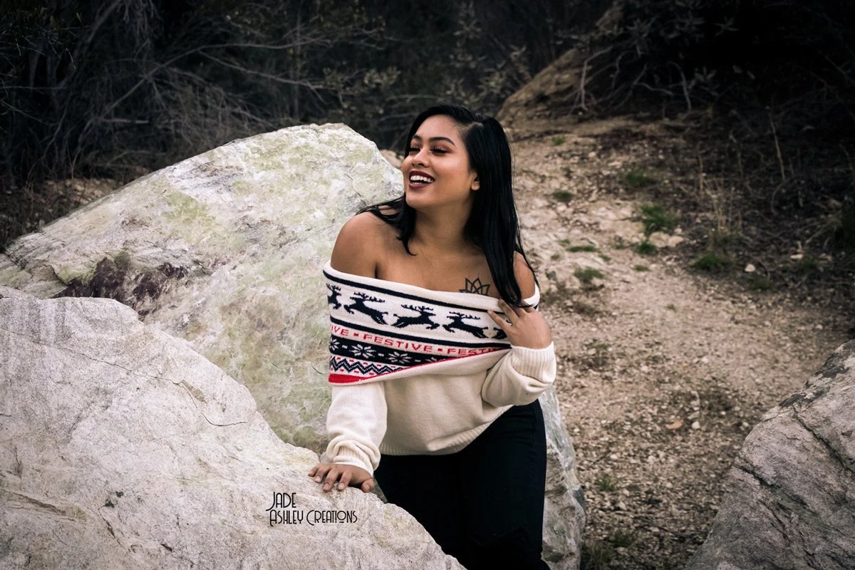 A smiling woman with dark hair and tattoo on shoulder, wearing an off-shoulder sweater with reindeer and holiday patterns, sitting on a large rock in an outdoor setting with dirt and sparse vegetation.