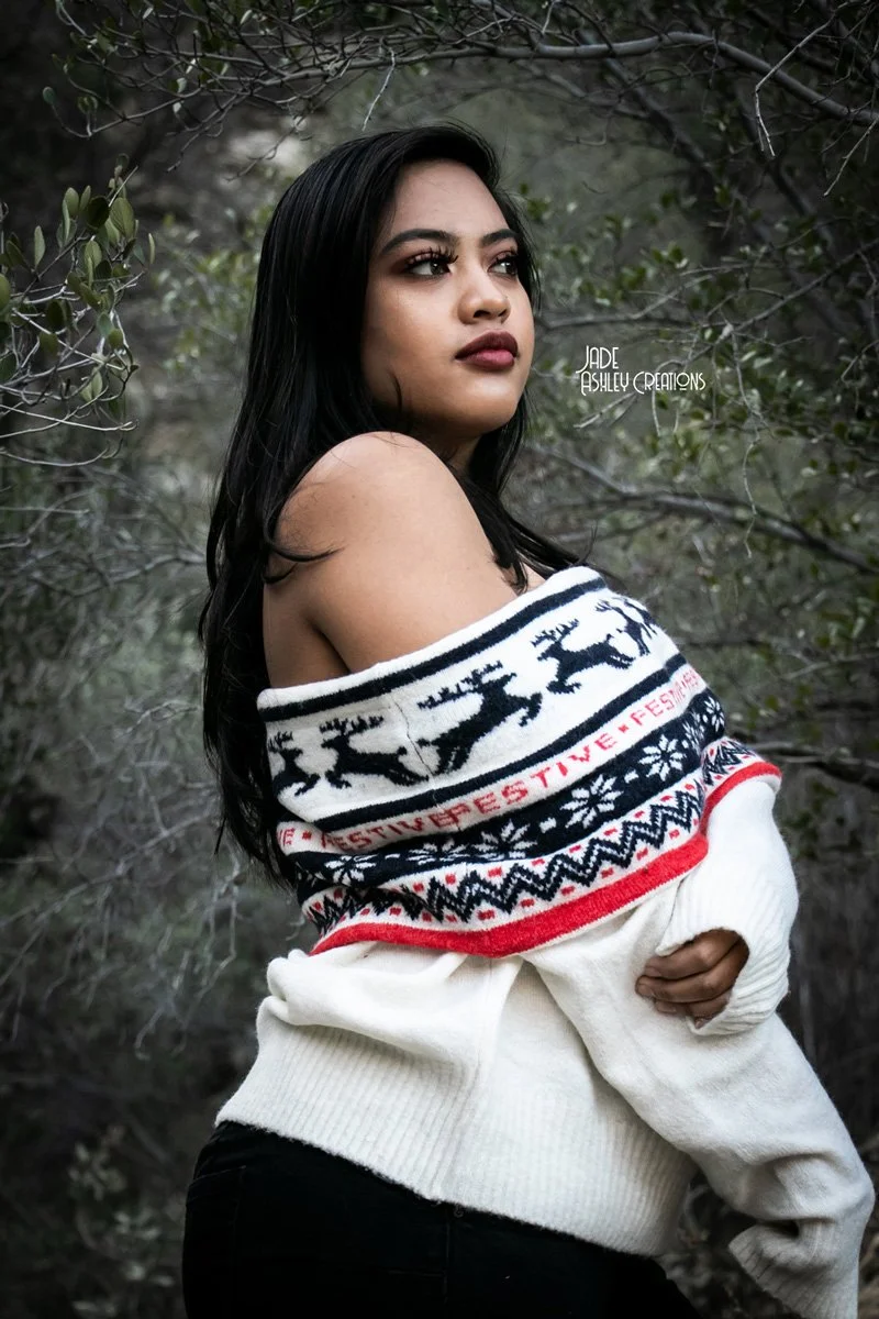 A woman with long dark hair and dark lipstick stands outdoors among thin, leafless branches and green foliage. She wears an off-the-shoulder sweater with a black, white, and red pattern depicting reindeer and snowflakes, and the words 'FESTIVE FESTIV