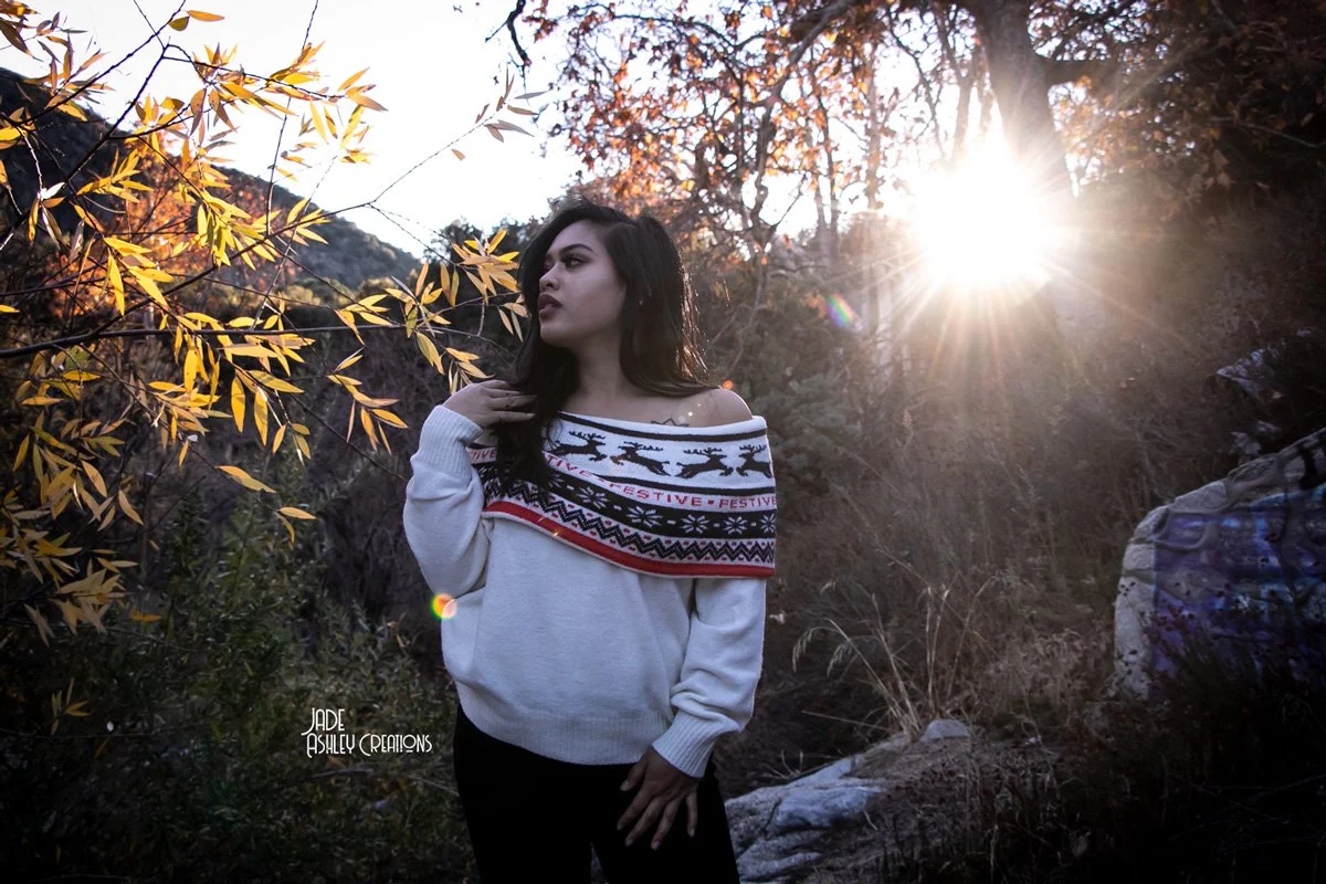 A woman standing outdoors among autumn trees with orange leaves, wearing a festive off-the-shoulder sweater, as the sun sets, creating a backlit scene.
