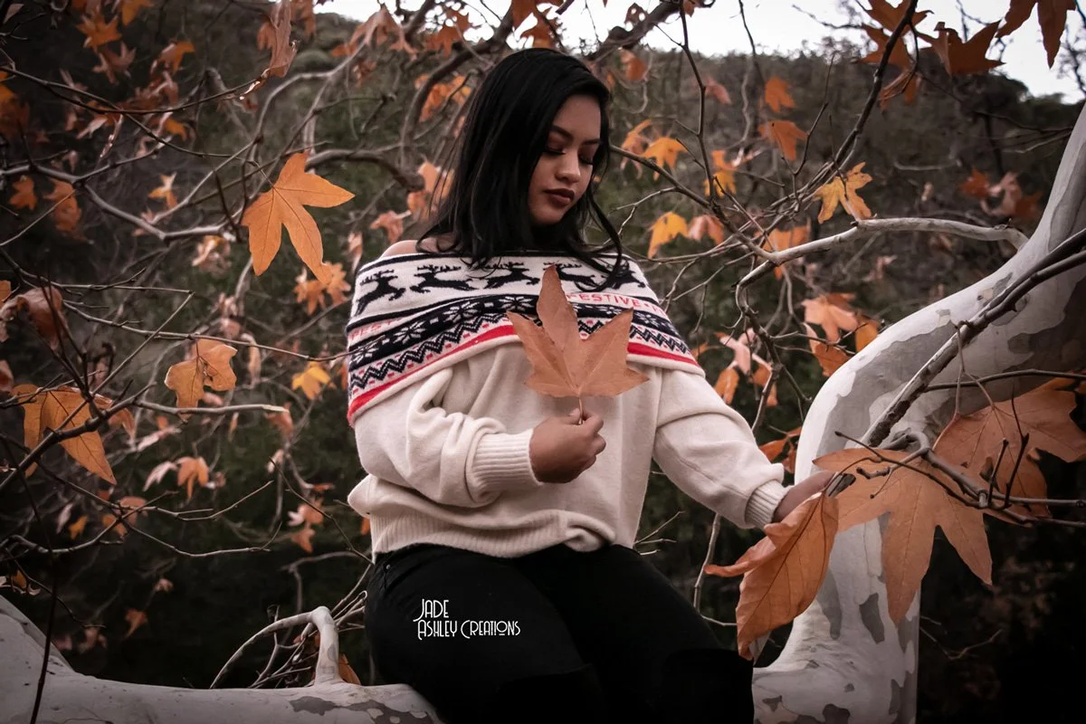 A young woman with black hair sitting on a tree branch surrounded by autumn leaves, holding a large maple leaf.