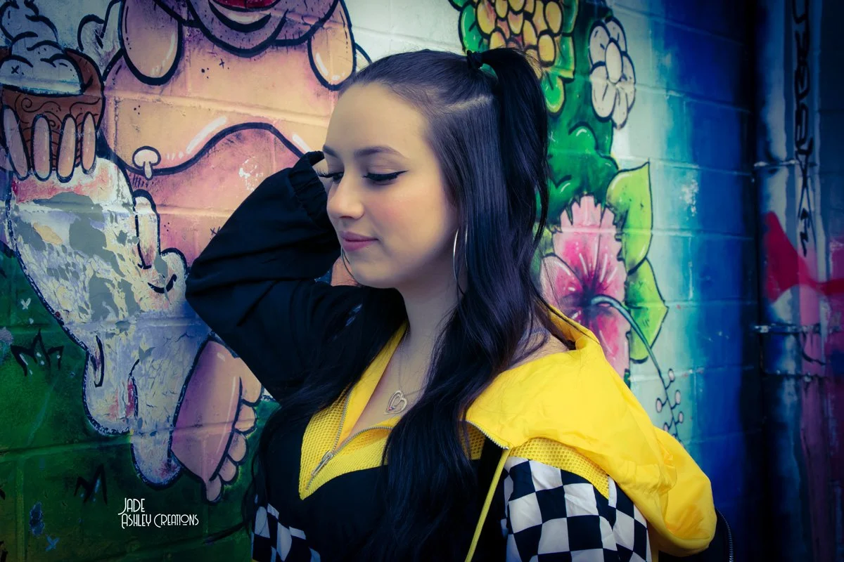 A young woman with long dark hair, partially tied, standing next to a colorful graffiti wall featuring a skull and floral designs, wearing a yellow and black jacket with checkered sleeves, and large hoop earrings.