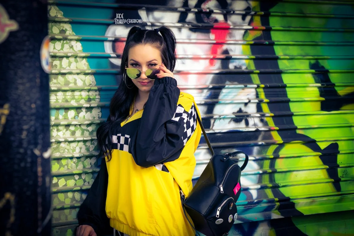 Young woman with dark hair in pigtails wearing sunglasses, black hoop earrings, and a yellow and black checkered jacket, standing in front of a colorful graffiti wall.