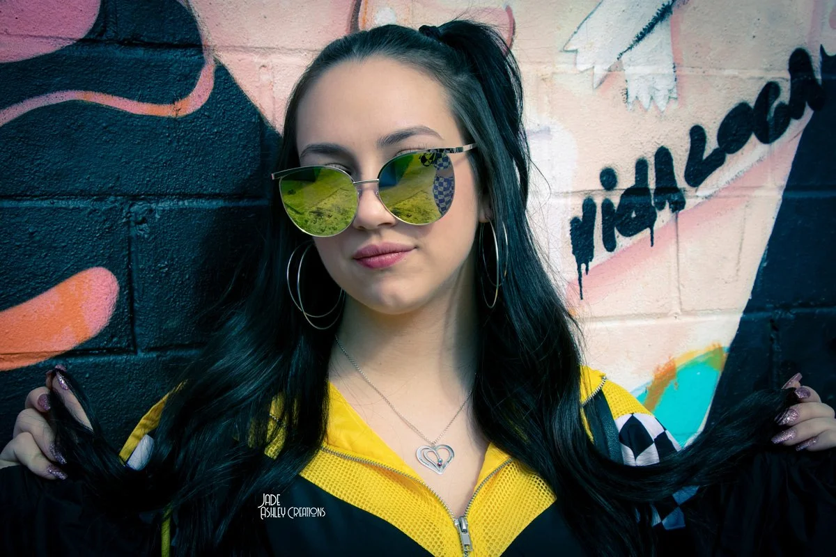 Young woman with long black hair wearing yellow sunglasses, hoop earrings, a necklace, and a yellow and black jacket, standing in front of a colorful graffiti wall.