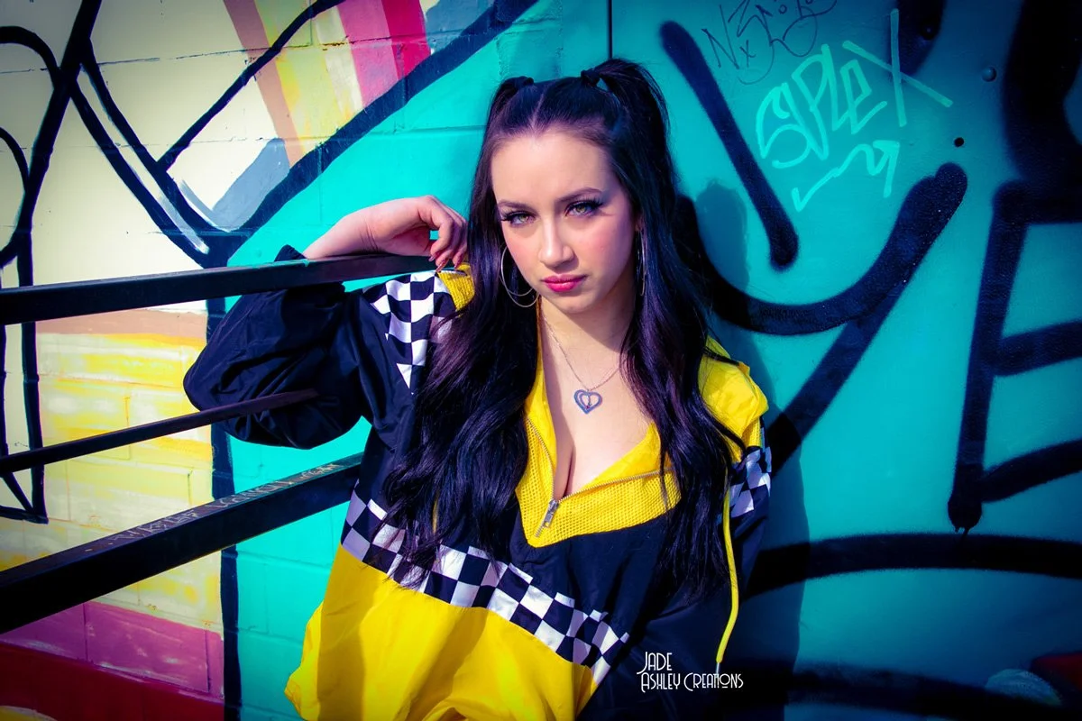 A young woman with long dark hair and hoop earrings poses against a vibrant graffiti wall, wearing a yellow and black checkered and checkered sports jacket.