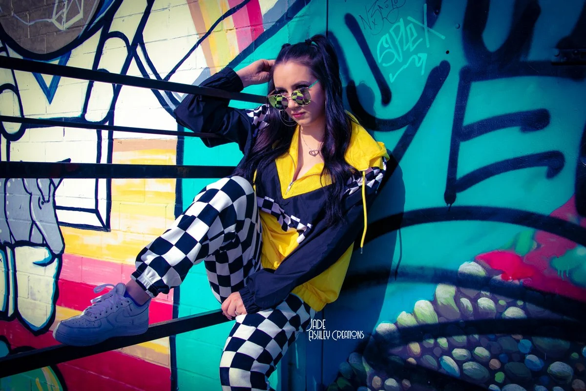 A young woman with long dark hair wearing colorful sunglasses, a yellow and black windbreaker, and black and white checkered pants, sitting against a vibrant graffiti wall.