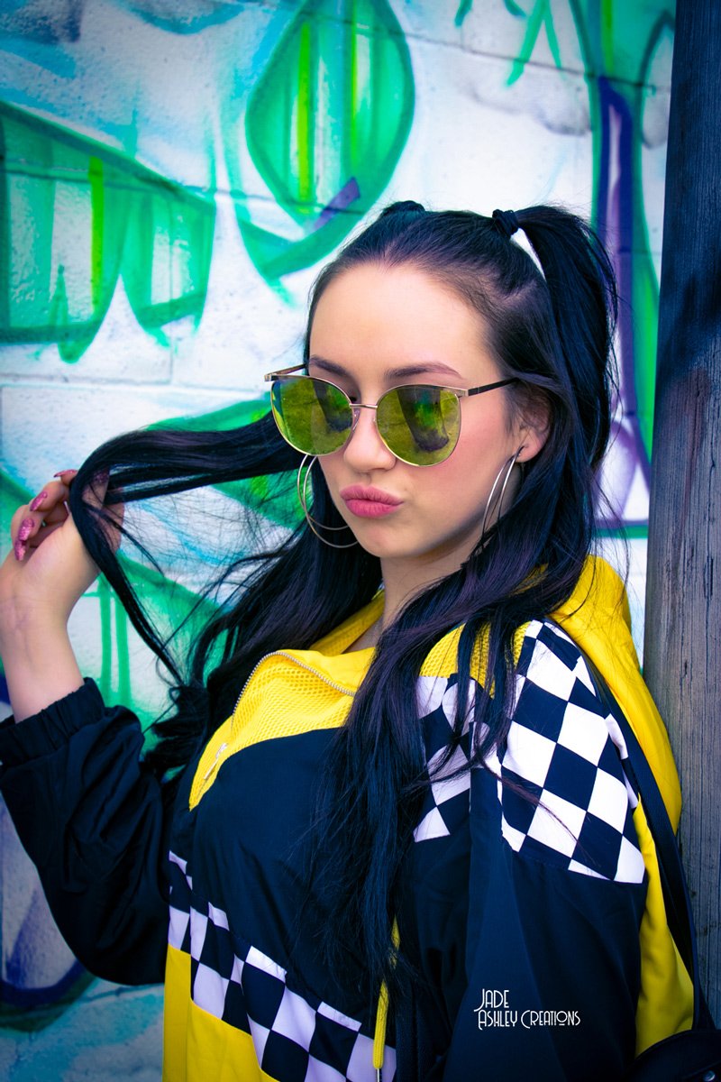 Young woman with black hair in a high ponytail wearing reflective green sunglasses, large hoop earrings, a yellow and black checkered jacket, against a graffiti wall background.