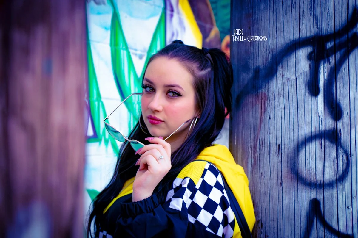 Young woman with dark hair and purple eyes holding sunglasses, leaning against a graffiti-covered wooden wall.