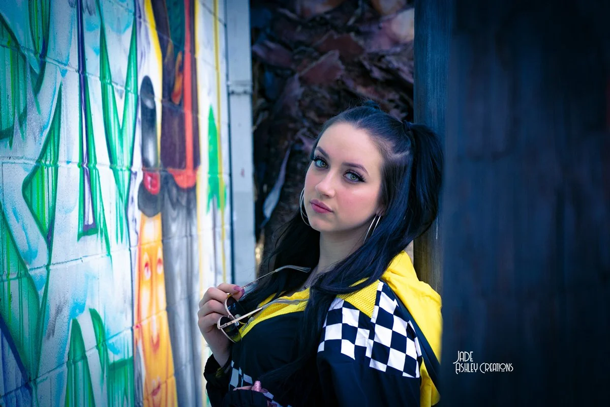 Young woman with black hair in pigtails, wearing hoop earrings and a black and white checkered jacket with yellow accents, stands next to a colorful graffiti wall, holding a pair of glasses.