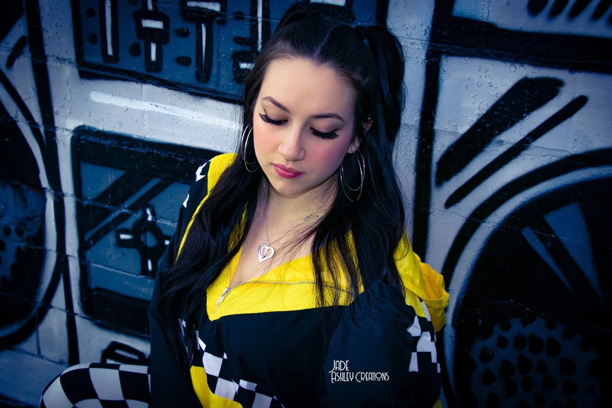 A young woman with long black hair, hoop earrings, and a necklace, standing against a graffiti-covered wall. She is wearing a yellow and black track jacket with a checkered pattern.
