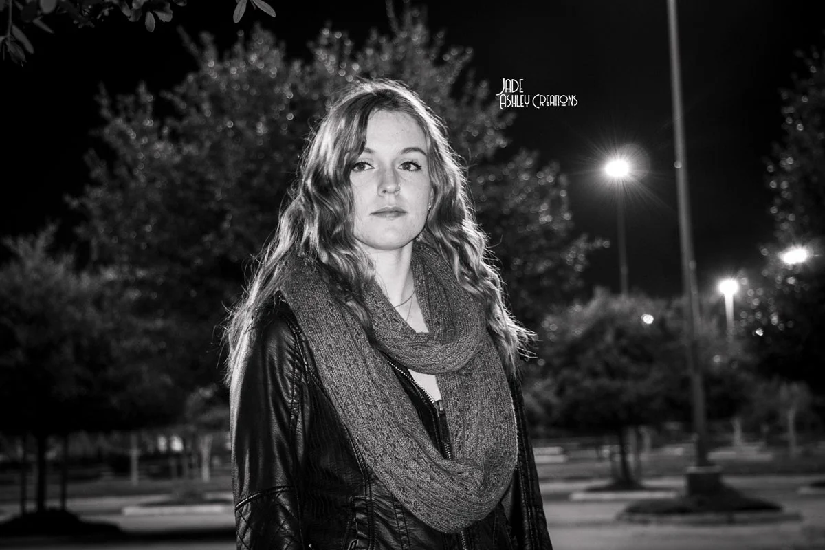 A young woman with wavy hair wearing a leather jacket and a scarf, standing outdoors at night with trees and streetlights in the background.
