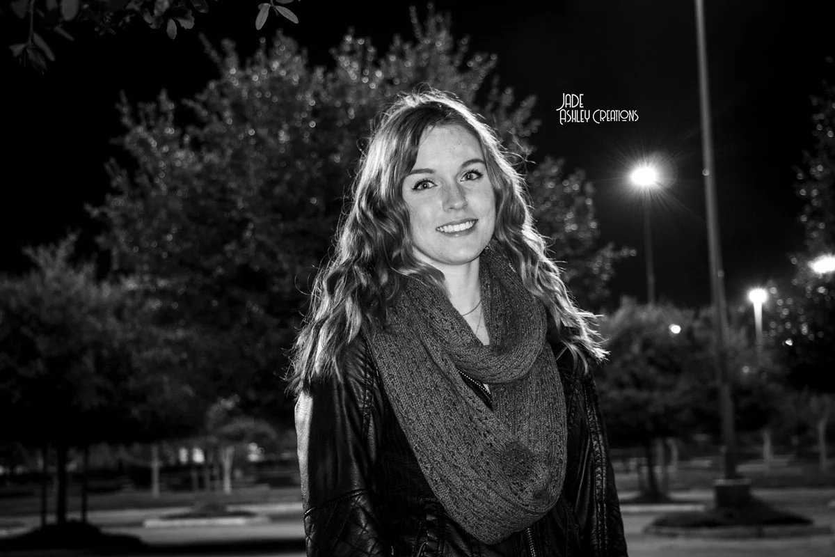 A young woman with wavy hair smiling outdoors at night, wearing a dark jacket and a large knit scarf, standing in front of trees and streetlights.