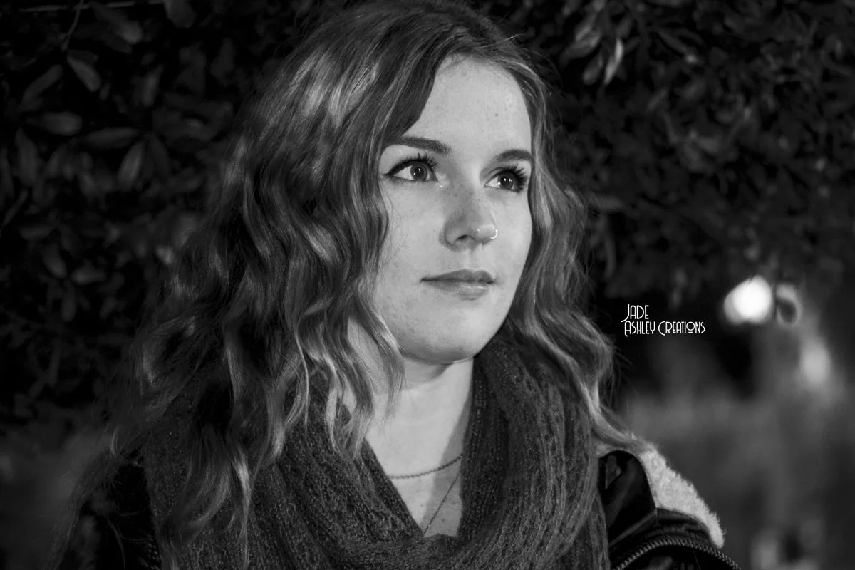 A young woman with wavy hair and freckles, wearing a scarf and jacket, looking off to the side outdoors at night.