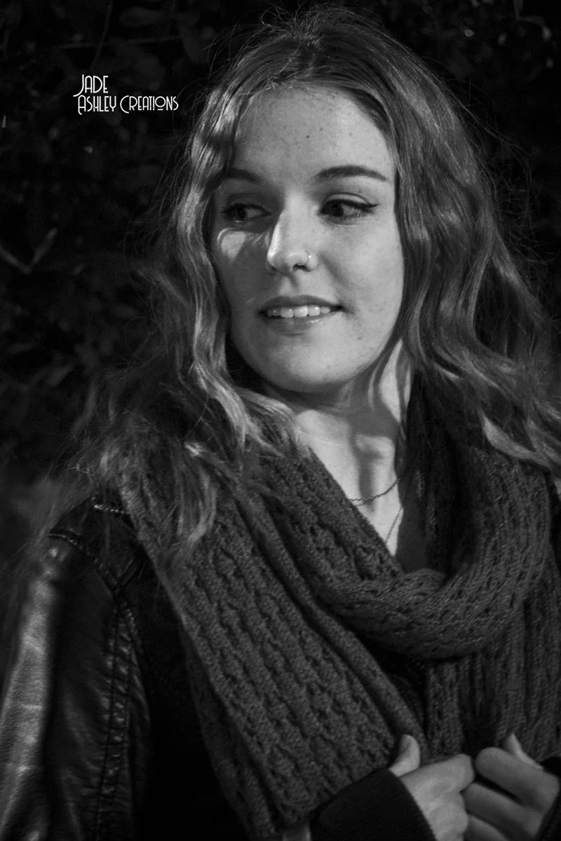 A young woman with wavy hair and a nose piercing, wearing a knit scarf and leather jacket, looking to her right outdoors at night.