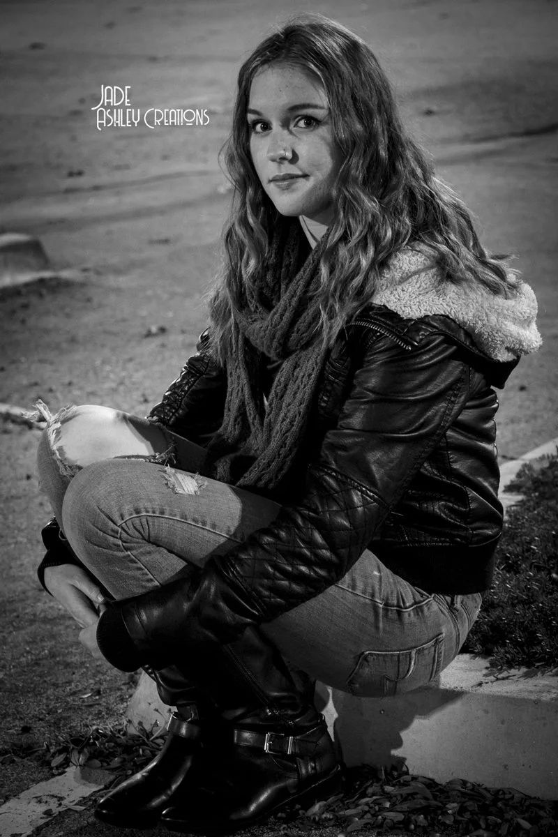 A young woman with long, wavy hair sitting on a curb outdoors at night, wearing a leather jacket with a fur-lined hood, a scarf, ripped jeans, and black boots.