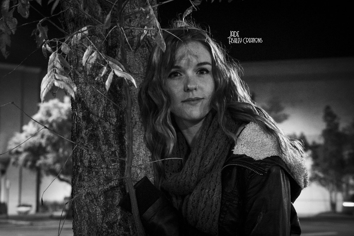 A young woman leaning against a tree at night, looking at the camera, with leaves and blurred background in a black-and-white photo.